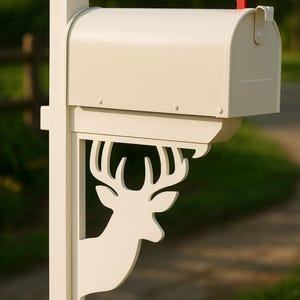 May include: Cream-colored mailbox with a red flag, mounted on a post. The post features a decorative deer head cutout. The mailbox is designed for outdoor use and is set against a blurred green background.