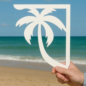 May include: A white wooden cutout of a palm tree, held in front of a beach scene with blue water and white sand.