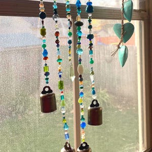 May include: A beaded wind chime with a wooden rod and hanging bronze bells. The strands feature a variety of colorful beads in shades of blue, green, yellow, and clear. Decorative heart-shaped elements are also visible.