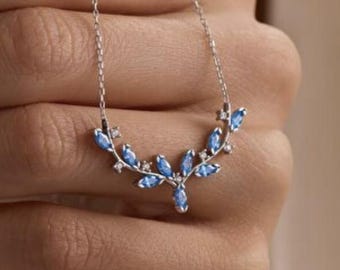 Puede incluir: Un collar de plata con un diseño curvo adornado con piedras preciosas azul claro. El collar se muestra, destacando sus intrincados detalles y la forma en que captura la luz. Las piedras están dispuestas en un patrón en forma de hoja.