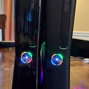 K&ouml;nnte beinhalten: Zwei vertikal nebeneinander stehende Xbox 360 Spielkonsolen. Die linke Konsole hat eine gl&auml;nzend schwarze Oberfl&auml;che, w&auml;hrend die rechte Konsole eine mattschwarze Oberfl&auml;che aufweist. Beide Ger&auml;te zeigen das Xbox 360 Logo und einen runden Einschaltknopf mit blau leuchtendem Licht.