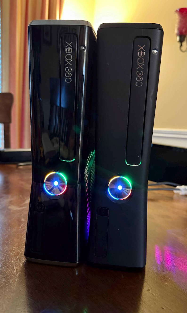 K&ouml;nnte beinhalten: Zwei vertikal nebeneinander stehende Xbox 360 Spielkonsolen. Die linke Konsole hat eine gl&auml;nzend schwarze Oberfl&auml;che, w&auml;hrend die rechte Konsole eine mattschwarze Oberfl&auml;che aufweist. Beide Ger&auml;te zeigen das Xbox 360 Logo und einen runden Einschaltknopf mit blau leuchtendem Licht.