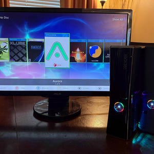 K&ouml;nnte beinhalten: Eine schwarze Xbox 360 Spielkonsole mit leuchtendem Power-Knopf steht neben einem Computermonitor, der die Aurora-Dashboard-Oberfl&auml;che anzeigt. Der Bildschirm zeigt verschiedene Anwendungssymbole sowie das gr&uuml;ne Aurora-Logo.