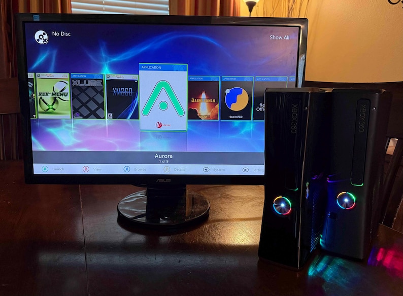 K&ouml;nnte beinhalten: Eine schwarze Xbox 360 Spielkonsole mit leuchtendem Power-Knopf steht neben einem Computermonitor, der die Aurora-Dashboard-Oberfl&auml;che anzeigt. Der Bildschirm zeigt verschiedene Anwendungssymbole sowie das gr&uuml;ne Aurora-Logo.