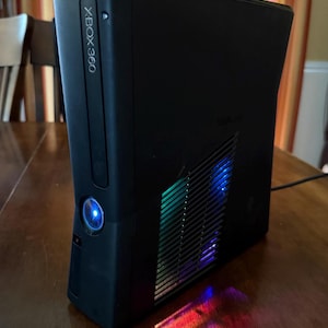 K&ouml;nnte beinhalten: Eine schwarze Xbox 360 Spielkonsole in vertikaler Ausrichtung. Die Vorderseite zeigt das Xbox 360 Logo und einen leuchtend blauen Power-Knopf. Das Seitenteil verf&uuml;gt &uuml;ber ein L&uuml;ftungsgitter, durch das mehrfarbige LED-Lichter hindurchscheinen.