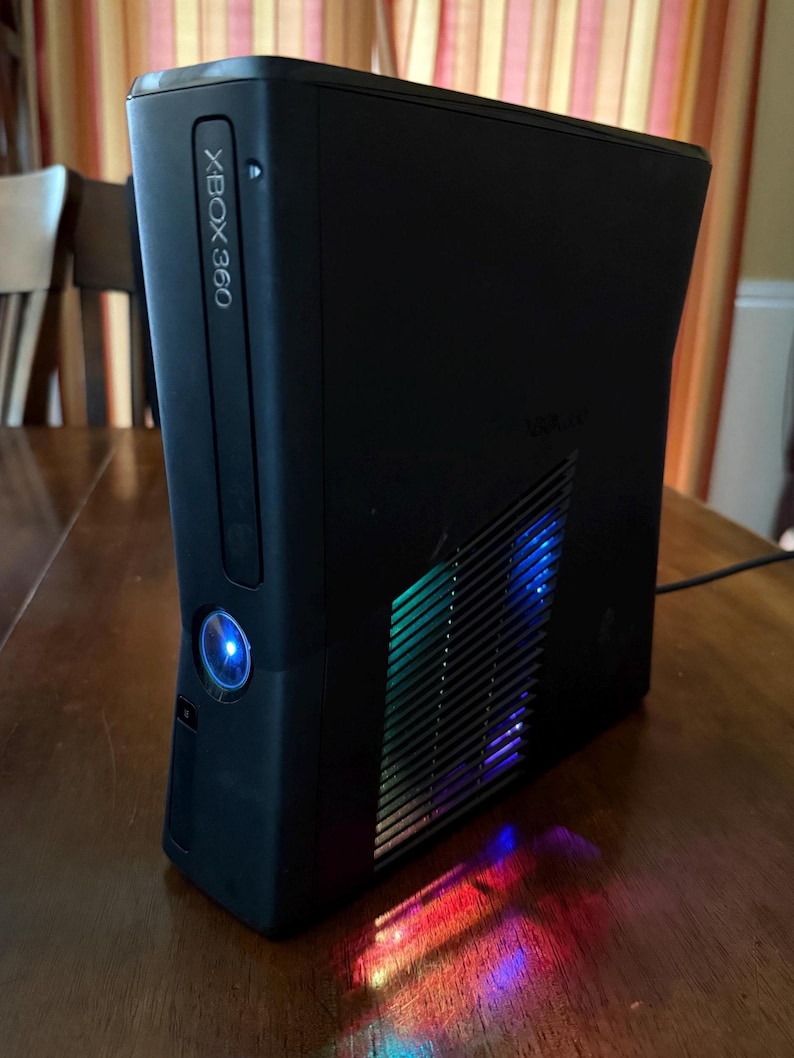 K&ouml;nnte beinhalten: Eine schwarze Xbox 360 Spielkonsole in vertikaler Ausrichtung. Die Vorderseite zeigt das Xbox 360 Logo und einen leuchtend blauen Power-Knopf. Das Seitenteil verf&uuml;gt &uuml;ber ein L&uuml;ftungsgitter, durch das mehrfarbige LED-Lichter hindurchscheinen.