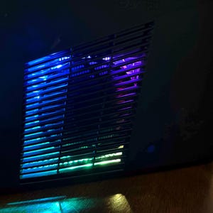 K&ouml;nnte beinhalten: Eine schwarze Spielkonsole mit einem schr&auml;gen, geschlitzten Seitenl&uuml;ftungsgitter. Das Innere des Gitters wird durch lebendige, wechselnde LED-Lichter in Blau-, Lila- und Gr&uuml;nt&ouml;nen beleuchtet, die einen leuchtenden Effekt durch das dunkle Kunststoffgitter erzeugen.