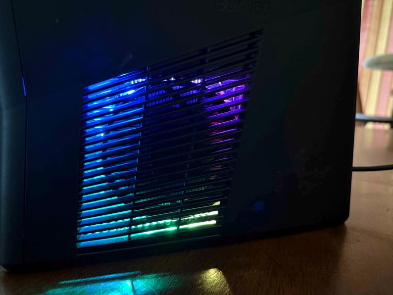 K&ouml;nnte beinhalten: Eine schwarze Spielkonsole mit einem schr&auml;gen, geschlitzten Seitenl&uuml;ftungsgitter. Das Innere des Gitters wird durch lebendige, wechselnde LED-Lichter in Blau-, Lila- und Gr&uuml;nt&ouml;nen beleuchtet, die einen leuchtenden Effekt durch das dunkle Kunststoffgitter erzeugen.