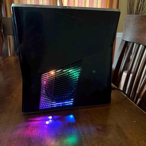 K&ouml;nnte beinhalten: Eine schwarze Spielkonsole mit gl&auml;nzender Oberfl&auml;che und einem zentralen L&uuml;ftungsgitter. Das Gitter strahlt lebendige, mehrfarbige LED-Lichter in Blau-, Gr&uuml;n- und Violettt&ouml;nen aus, die einen farbigen Schein auf die Oberfl&auml;che darunter werfen.