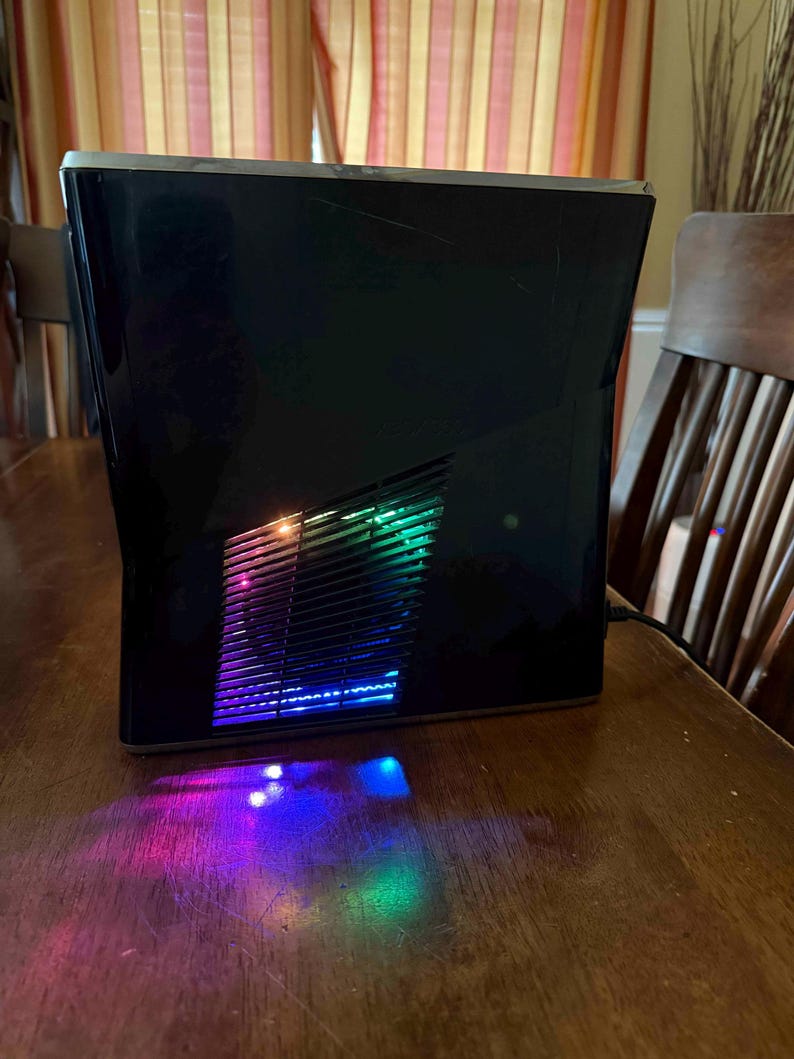K&ouml;nnte beinhalten: Eine schwarze Spielkonsole mit gl&auml;nzender Oberfl&auml;che und einem zentralen L&uuml;ftungsgitter. Das Gitter strahlt lebendige, mehrfarbige LED-Lichter in Blau-, Gr&uuml;n- und Violettt&ouml;nen aus, die einen farbigen Schein auf die Oberfl&auml;che darunter werfen.