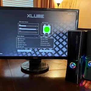 K&ouml;nnte beinhalten: Ein ASUS-Computermonitor zeigt die XLUME-Softwareoberfl&auml;che mit RGB-Farbeinstellungen und LED-Vorschau, platziert neben zwei schwarzen Xbox 360-Spielkonsolen mit beleuchteten, mehrfarbigen kreisf&ouml;rmigen Ein-Aus-Schaltern an der Vorderseite.