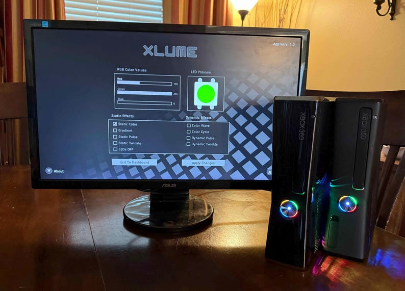 K&ouml;nnte beinhalten: Ein ASUS-Computermonitor zeigt die XLUME-Softwareoberfl&auml;che mit RGB-Farbeinstellungen und LED-Vorschau, platziert neben zwei schwarzen Xbox 360-Spielkonsolen mit beleuchteten, mehrfarbigen kreisf&ouml;rmigen Ein-Aus-Schaltern an der Vorderseite.
