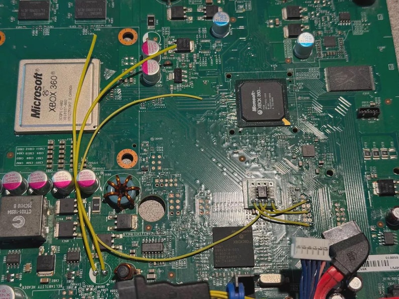 K&ouml;nnte beinhalten: Ein gr&uuml;nes Xbox 360-Mainboard mit verschiedenen elektronischen Komponenten, darunter ein Hauptprozessor mit Microsoft-Branding, silberne Kondensatoren und mehrere d&uuml;nne gelbe Dr&auml;hte, die auf die Oberfl&auml;che der Platine gel&ouml;tet sind.