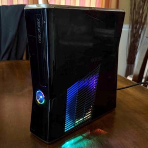 K&ouml;nnte beinhalten: Eine gl&auml;nzend schwarze Xbox 360 Spielkonsole in vertikaler Ausrichtung. Die Vorderseite zeigt das Xbox 360 Logo und einen kreisf&ouml;rmigen Power-Knopf mit blauer Beleuchtung. Ein seitliches Gitter strahlt ein blaues und violettes Licht aus.
