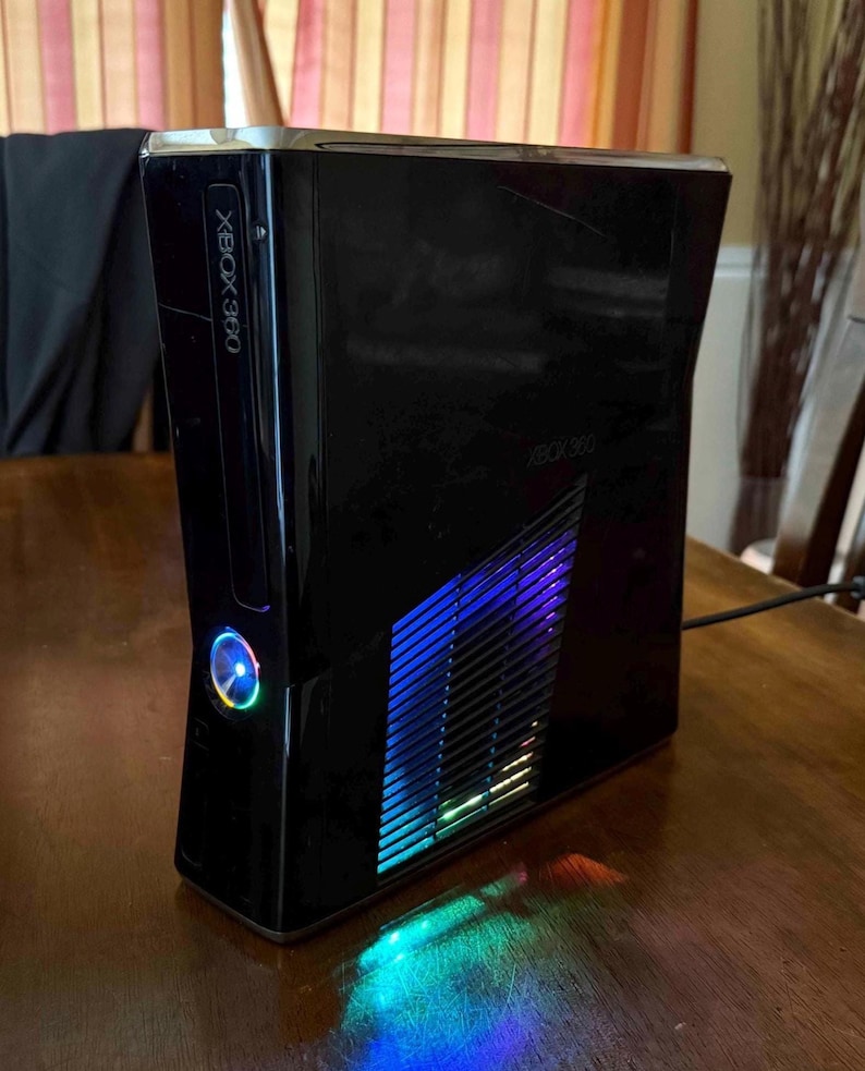 K&ouml;nnte beinhalten: Eine gl&auml;nzend schwarze Xbox 360 Spielkonsole in vertikaler Ausrichtung. Die Vorderseite zeigt das Xbox 360 Logo und einen kreisf&ouml;rmigen Power-Knopf mit blauer Beleuchtung. Ein seitliches Gitter strahlt ein blaues und violettes Licht aus.