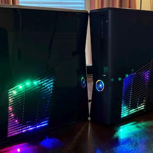 K&ouml;nnte beinhalten: Zwei schwarze Xbox 360 Spielkonsolen, die vertikal aufgestellt sind. Beide Einheiten verf&uuml;gen &uuml;ber eine gl&auml;nzende Oberfl&auml;che und ein seitliches L&uuml;ftungsgitter, das von internen, mehrfarbigen LED-Lichtern beleuchtet wird. Die Power-Tasten leuchten blau.
