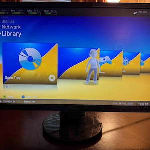 K&ouml;nnte beinhalten: Ein Computermonitor zeigt eine digitale Benutzeroberfl&auml;che mit einem blau-gelben Farbverlaufshintergrund. Der Bildschirm zeigt ein Men&uuml; mit dem Titel Library mit Symbolen f&uuml;r Open Tray, Games und andere Optionen sowie eine wei&szlig;e Avatarfigur.