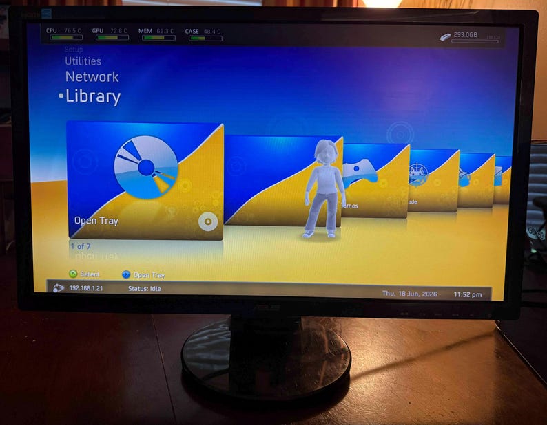 K&ouml;nnte beinhalten: Ein Computermonitor zeigt eine digitale Benutzeroberfl&auml;che mit einem blau-gelben Farbverlaufshintergrund. Der Bildschirm zeigt ein Men&uuml; mit dem Titel Library mit Symbolen f&uuml;r Open Tray, Games und andere Optionen sowie eine wei&szlig;e Avatarfigur.