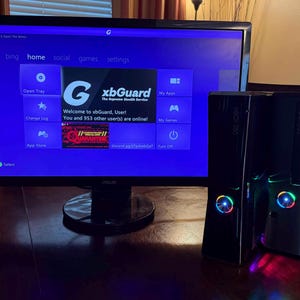 K&ouml;nnte beinhalten: Zwei schwarze Xbox 360 Spielkonsolen mit beleuchteten Power-Tasten stehen neben einem Asus-Monitor, der eine violette Dashboard-Oberfl&auml;che anzeigt. Der Bildschirm zeigt den Text xbGuard, The Supreme Stealth Service und ein Men&uuml; mit Optionen.