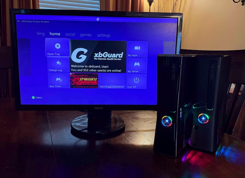 K&ouml;nnte beinhalten: Zwei schwarze Xbox 360 Spielkonsolen mit beleuchteten Power-Tasten stehen neben einem Asus-Monitor, der eine violette Dashboard-Oberfl&auml;che anzeigt. Der Bildschirm zeigt den Text xbGuard, The Supreme Stealth Service und ein Men&uuml; mit Optionen.