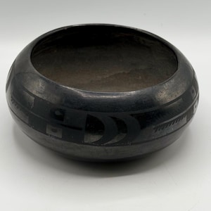 Puede incluir: Un cuenco de cerámica redondo gris oscuro con una abertura ancha. El cuenco presenta un diseño geométrico negro alrededor del exterior. El diseño incluye formas rectangulares y curvas. El cuenco está sobre una superficie blanca.