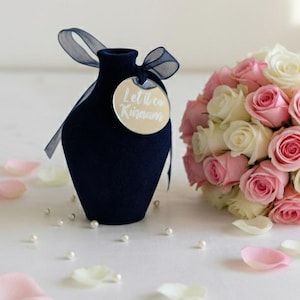 Puede incluir: Un jarrón azul marino con una cinta y una etiqueta que dice "Let it be Kiranam" junto a un ramo de rosas rosas y blancas. Pétalos y perlas dispersos complementan el arreglo floral.