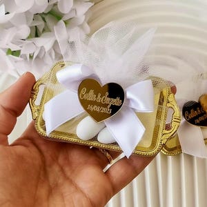 Pode incluir: Uma bandeja dourada e ornamentada cheia de doces brancos, embrulhada em tule branco e amarrada com uma fita branca. Uma etiqueta dourada em forma de coração com o texto "Collin & Suzette 16/08/2023" está presa à fita.