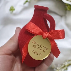 Puede incluir: Una caja de regalo roja en forma de jarrón con una etiqueta dorada y una cinta roja. La etiqueta tiene texto en idioma extranjero y la fecha 7.06.25. Rosas blancas y vegetación en el fondo.