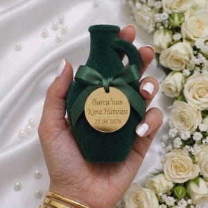 Può includere: Un vaso decorativo verde scuro, dalla texture vellutata, con manico, legato con un nastro verde e un'etichetta dorata. L'etichetta ha del testo sopra. Il vaso è tenuto in mano, con braccialetti d'oro al polso.