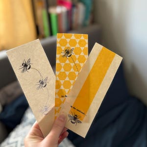 Könnte beinhalten: Drei rechteckige Lesezeichen mit Bienenillustrationen. Eines zeigt fliegende Bienen, ein anderes hat ein gelbes Wabenmuster und das dritte einen gelben Streifen. Der Text "to be continued..." erscheint auf zwei der Lesezeichen.