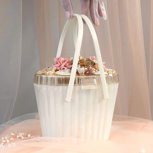 Può includere: Una borsa bianca in stile vintage con un corpo trasparente e scanalato e un bordo dorato. La borsa è adornata con fiori rosa, perle e conchiglie. Ha manici bianchi ed è tenuta da una mano che indossa un anello d'oro.