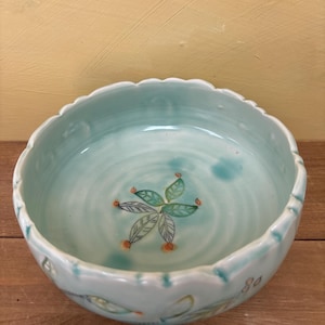 May include: A round ceramic bowl with a scalloped rim, glazed in a light seafoam green color. The interior features a hand-painted botanical motif of green leaves and small orange dots at the center, with a rippled texture throughout the surface.