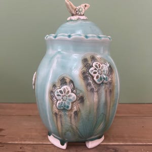 May include: A turquoise ceramic jar with a lid, featuring floral designs and a bird-shaped finial. The jar has a rounded body, three small feet, and a ruffled edge on the lid. The floral patterns are in shades of white, gray, and brown.