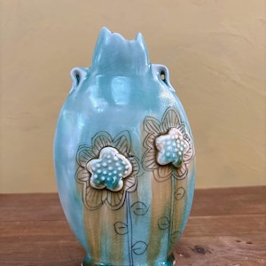 May include: A light blue ceramic vase with a crackle glaze finish, featuring two raised, textured white and green flower appliqués on the front. The vase has a scalloped base, two small side handles, and hand-drawn black floral stems.