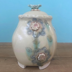 May include: A cream-colored ceramic jar with a lid, adorned with blue floral designs and green leaves. The jar has a small bird-shaped handle on the lid and sits on three small feet. The jar is approximately 6 inches tall.