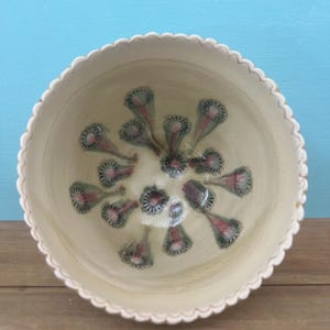 May include: Cream-colored ceramic bowl with a scalloped edge. The interior features a repeating pattern of stylized floral designs in shades of pink, green, and gray. The bowl is set against a light blue background.