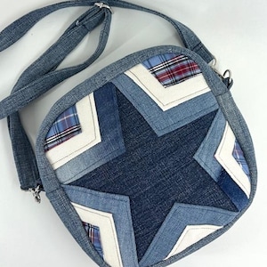 Op de afbeelding: Een denim crossbody tas met een sterontwerp. De tas heeft een patchwork ontwerp met een grote ster in het midden, gemaakt van verschillende tinten blauwe denim. Accentueren met witte stof en geruite inzetstukken. De verstelbare riem is ook van denim.