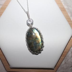 May include: A silver-toned pendant necklace featuring a large, oval-shaped labradorite gemstone with iridescent blue and gold flashes. The stone is encased in a decorative wire-wrapped setting, topped with a small, clear, faceted accent stone.