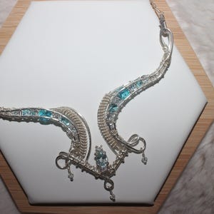 May include: A silver wire-wrapped necklace featuring a curved, asymmetrical design with embedded light blue and clear faceted stones. The metalwork consists of intricate coiled silver wire, with small dangling accents at the bottom center.