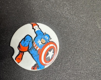 May include: A white car coaster featuring a comic book illustration of Captain America. The superhero is depicted in his classic blue, red, and white costume, holding his shield with a star emblem. The coaster is round and has a small notch.