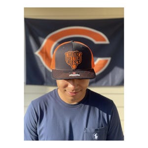 May include: A dark gray and orange baseball cap featuring a bear-shaped patch on the front panel with the text GOOD BETTER REST printed in orange. The cap has a structured crown, a flat brim, and a small rectangular sticker on the brim's center.