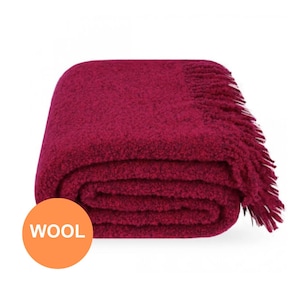 Peut inclure: Une couverture en laine pliée, de couleur bordeaux, avec une bordure à franges. La couverture est de couleur unie et semble douce et moelleuse. Le mot "WOOL" est imprimé sur un cercle orange en bas à gauche.