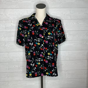 May include: Black short-sleeved button-up shirt with a colorful floral pattern. The shirt features a classic collar and white buttons. The floral design includes red, pink, yellow, and purple flowers with white vines.