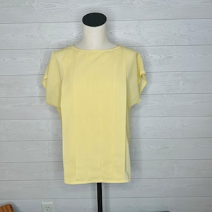 May include: A pale yellow short-sleeved blouse with vertical pleats down the front. The blouse is displayed on a mannequin with a black neck and stand, set against a white wood-paneled background. The top has a round neckline.