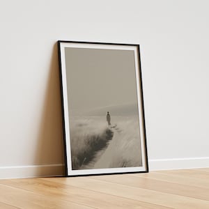 Könnte beinhalten: Gerahmter Kunstdruck in Schwarzweiß, der eine Person zeigt, die auf einem Weg durch hohes Gras geht. Das Kunstwerk ist vor einem gedämpften Himmel platziert. Der schwarz gerahmte Druck lehnt an einer weißen Wand auf einem Holzboden.