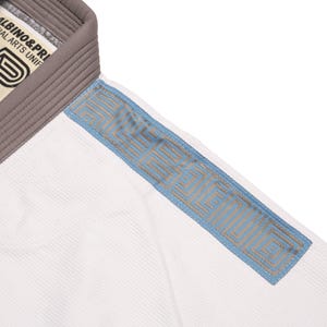 May include: A white martial arts gi top featuring a thick grey collar and a light blue shoulder panel with a repeating geometric pattern. The interior neck label displays the text ALBINO & PRETO MARTIAL ARTS UNIFORMS with a logo graphic.
