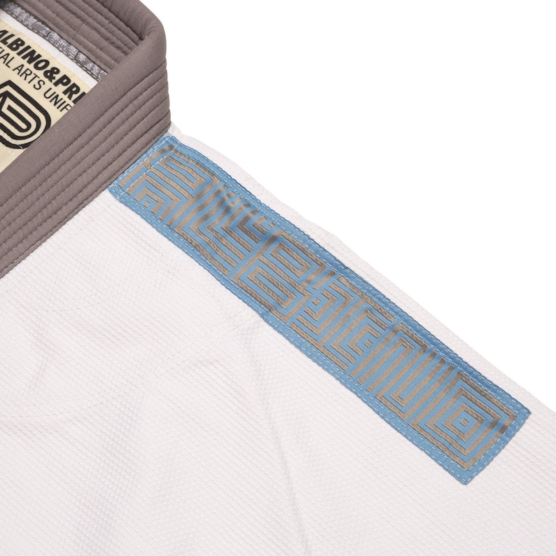 May include: A white martial arts gi top featuring a thick grey collar and a light blue shoulder panel with a repeating geometric pattern. The interior neck label displays the text ALBINO & PRETO MARTIAL ARTS UNIFORMS with a logo graphic.