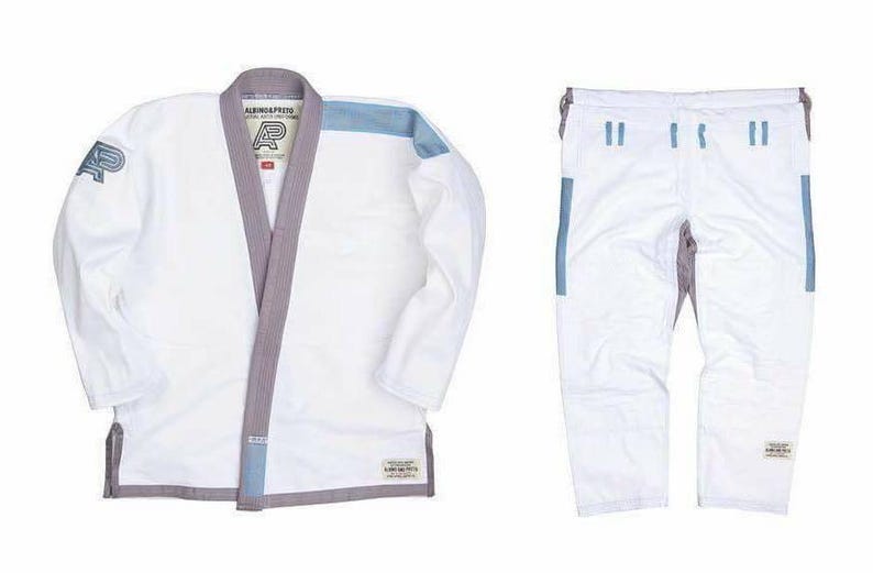 May include: A white Brazilian Jiu-Jitsu gi set consisting of a jacket and pants. The jacket features a grey collar, light blue shoulder accents, and the text Albino & Preto with an AP logo. The pants are white with grey and light blue detailing.