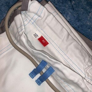 May include: A white cotton garment featuring light blue stitching, a beige drawstring cord, and two light blue fabric loops. A small white tag displays the text 100% COTTON MADE IN PAKISTAN, and a red rectangular tag features the white text A1.