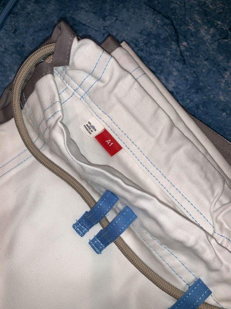 May include: A white cotton garment featuring light blue stitching, a beige drawstring cord, and two light blue fabric loops. A small white tag displays the text 100% COTTON MADE IN PAKISTAN, and a red rectangular tag features the white text A1.